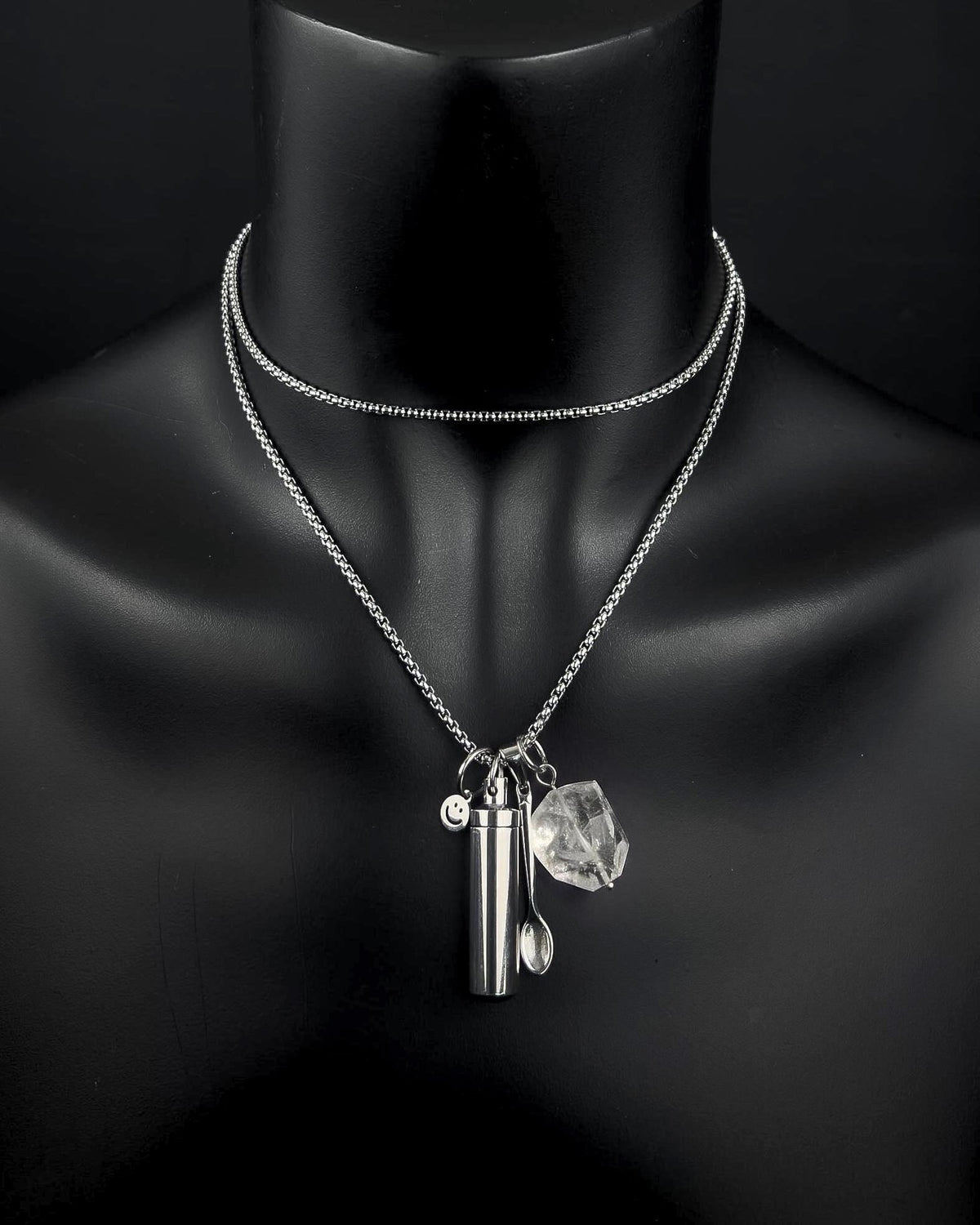 A silver-toned double-layered chain necklace featuring three hanging charms: a small cylindrical metal capsule, a tiny spoon, and a clear, faceted quartz stone. A small circular charm with a smiley face is also attached.