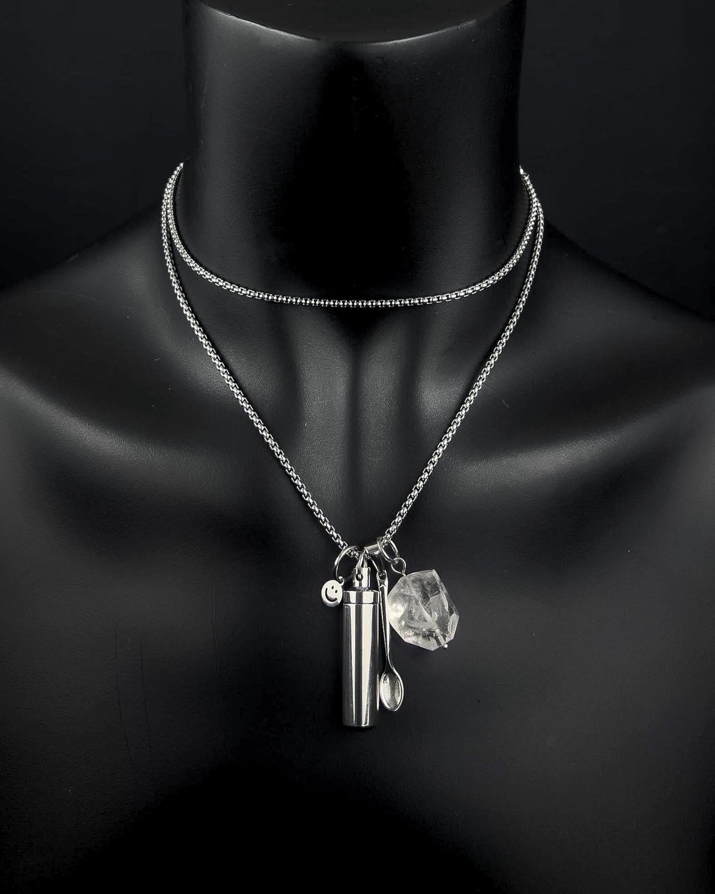 A silver-toned double-layered chain necklace featuring three hanging charms: a small cylindrical metal capsule, a tiny spoon, and a clear, faceted quartz stone. A small circular charm with a smiley face is also attached.
