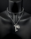 A silver-toned double-layered chain necklace featuring three hanging charms: a small cylindrical metal capsule, a tiny spoon, and a clear, faceted quartz stone. A small circular charm with a smiley face is also attached.