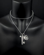A silver-toned double-layered chain necklace featuring three hanging charms: a small cylindrical metal capsule, a tiny spoon, and a clear, faceted quartz stone. A small circular charm with a smiley face is also attached.