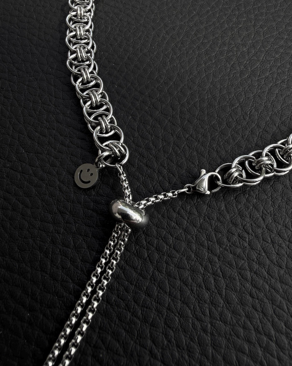 A silver-coloured stainless steel chain necklace featuring a complex interlocking circular link pattern. The necklace includes a sliding adjustable bead mechanism with two thin box chains hanging down and a small circular charm with a smiley face icon.