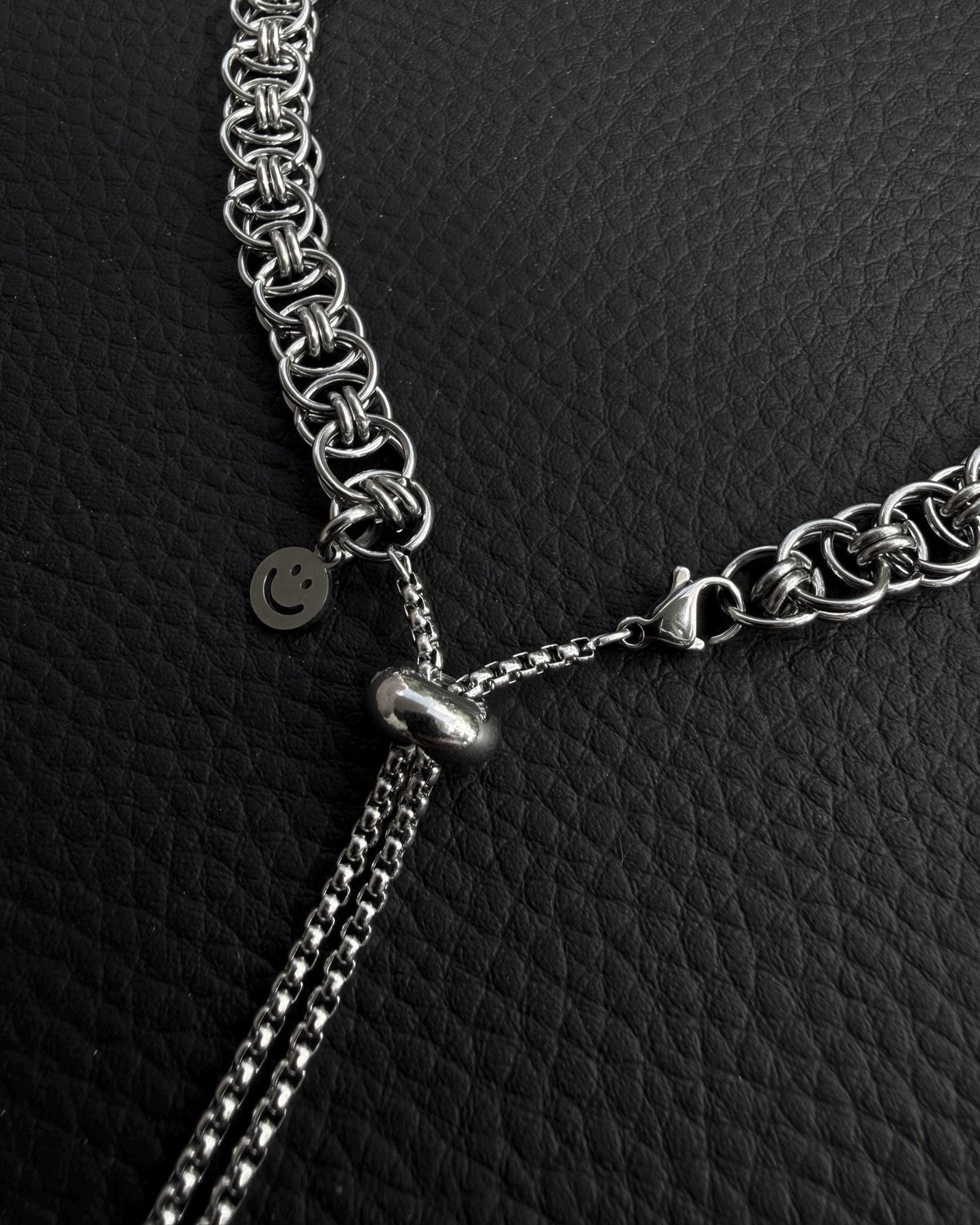 A silver-coloured stainless steel chain necklace featuring a complex interlocking circular link pattern. The necklace includes a sliding adjustable bead mechanism with two thin box chains hanging down and a small circular charm with a smiley face icon.