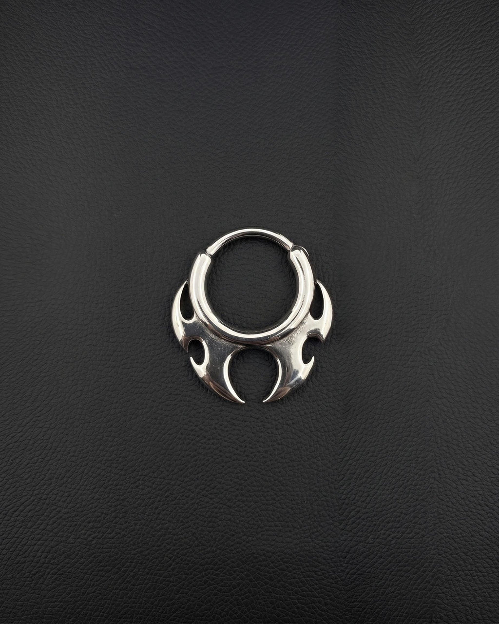 Handmade stainless steel tribal hoop earring with a spiky symmetrical design on a dark background designed as alternative rave jewelry. A silver-toned circular septum ring featuring a decorative, symmetrical tribal-inspired design with pointed, flame-like protrusions extending from the bottom half of the hoop. The metal has a polished, reflective finish. cyber sigilism style