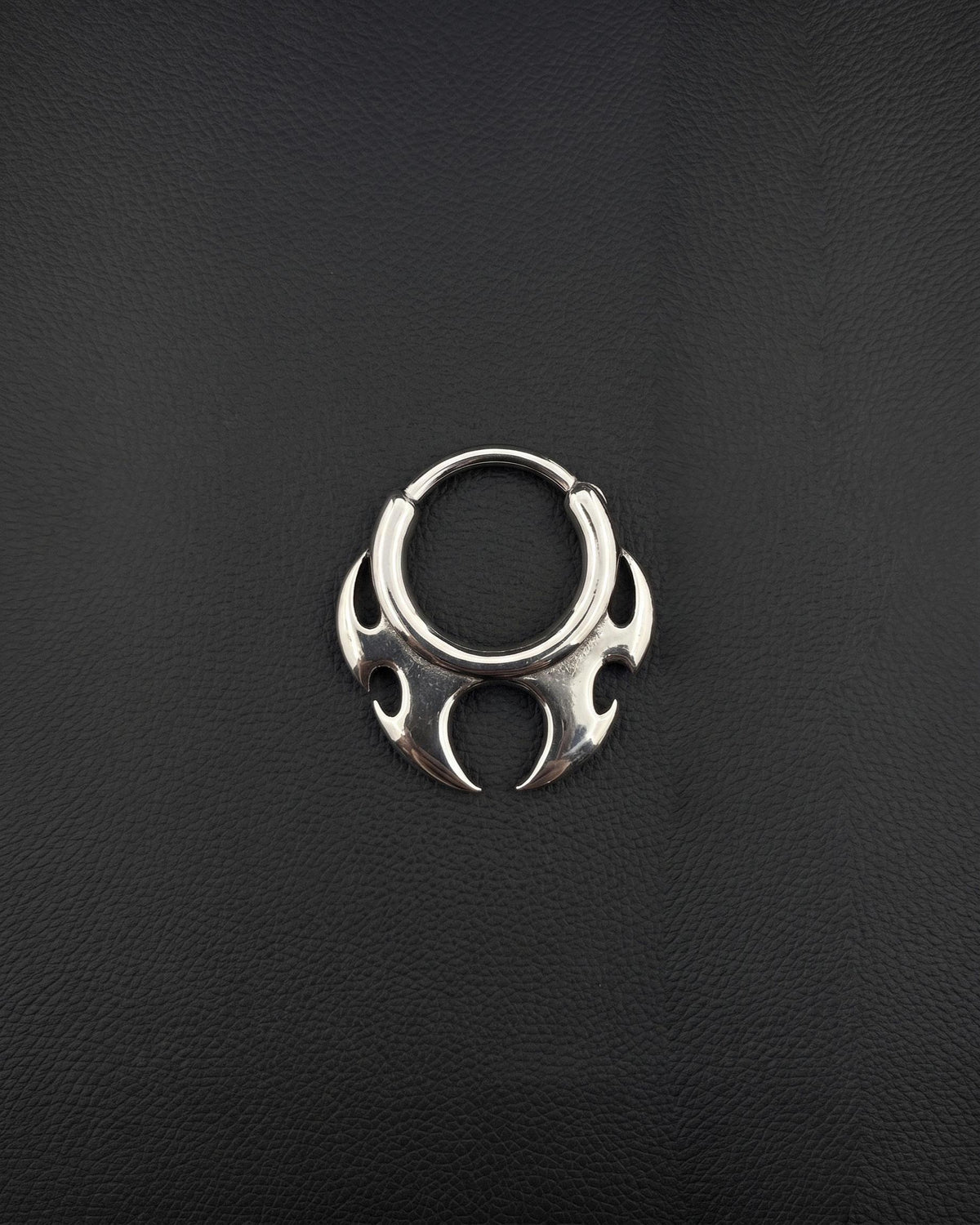 Handmade stainless steel tribal hoop earring with a spiky symmetrical design on a dark background designed as alternative rave jewelry. A silver-toned circular septum ring featuring a decorative, symmetrical tribal-inspired design with pointed, flame-like protrusions extending from the bottom half of the hoop. The metal has a polished, reflective finish. cyber sigilism style