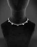 A silver stainless steel chain necklace featuring a series of evenly spaced, polished heart-shaped charms integrated into a complex, woven chainmaille metal link design. The necklace rests symmetrically, showcasing a repeating pattern of hearts and textured links.