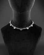 A silver stainless steel chain necklace featuring a series of evenly spaced, polished heart-shaped charms integrated into a complex, woven chainmaille metal link design. The necklace rests symmetrically, showcasing a repeating pattern of hearts and textured links.