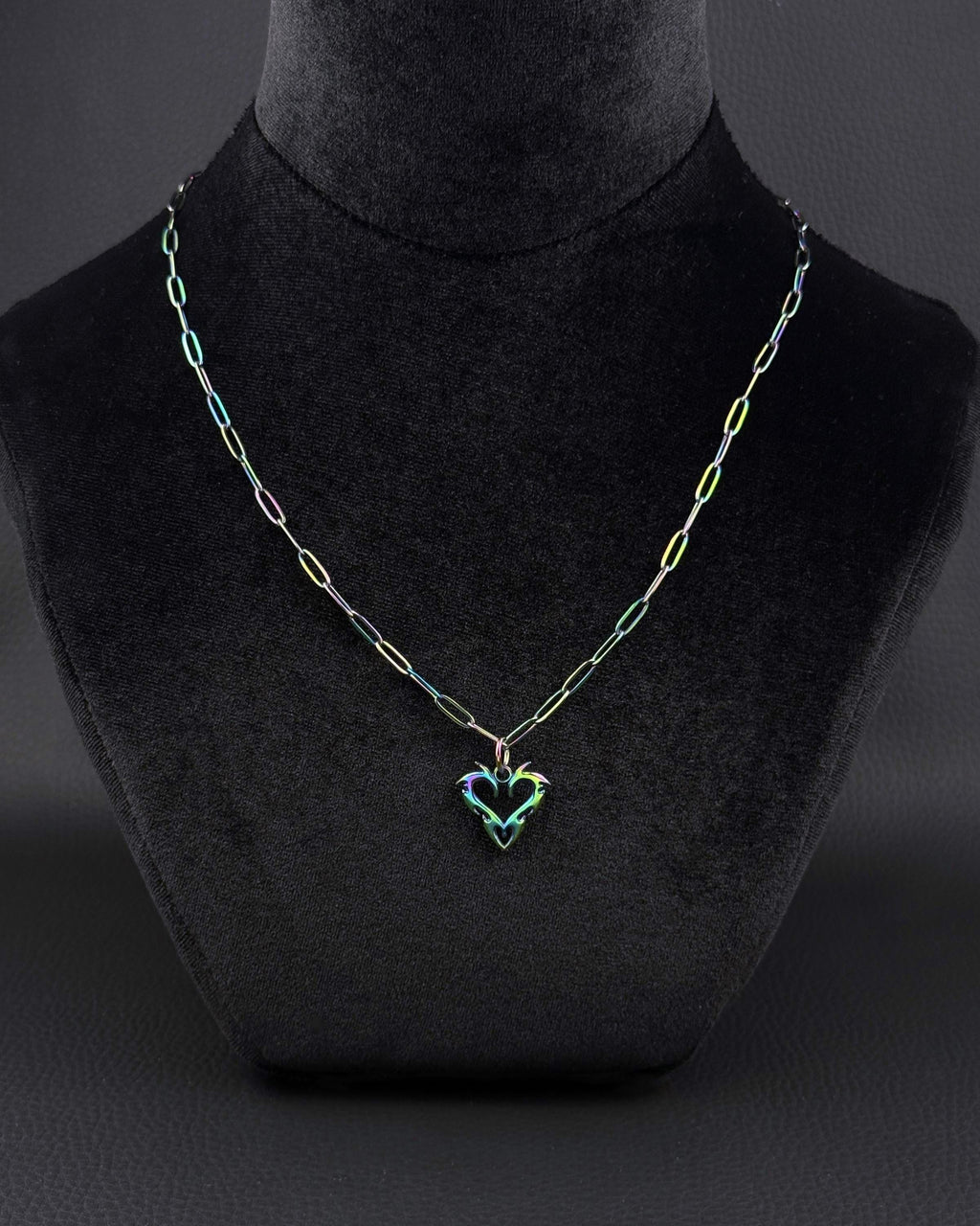 An iridescent oil-slick coloured, rainbow, stainless steel necklace featuring a paperclip style chain and a central heart-shaped pendant. The metal finish displays shifting hues of blue, green, and purple across the entire surface of the chain and the heart charm.