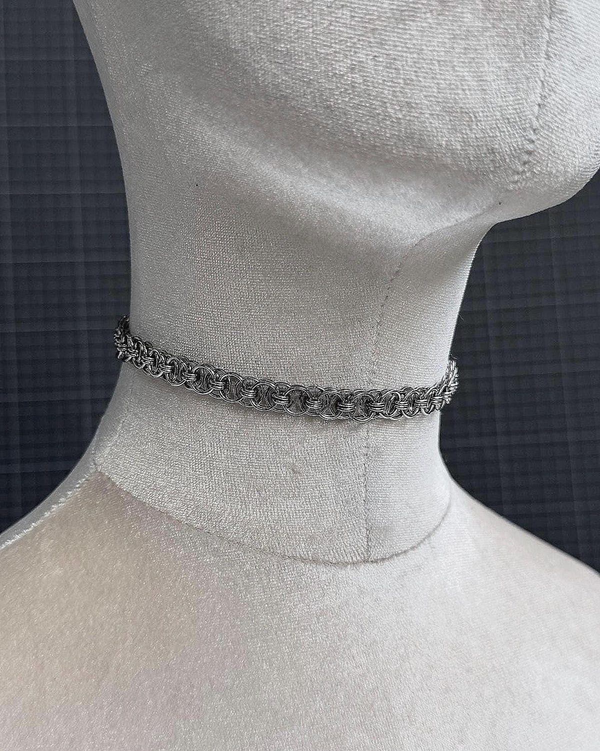 A silver-toned stainless steel chainmail tattoo choker necklace featuring a repeating pattern of interlocking metal rings. The necklace is constructed from small, circular links that create a textured, woven appearance around the neck of the display form.