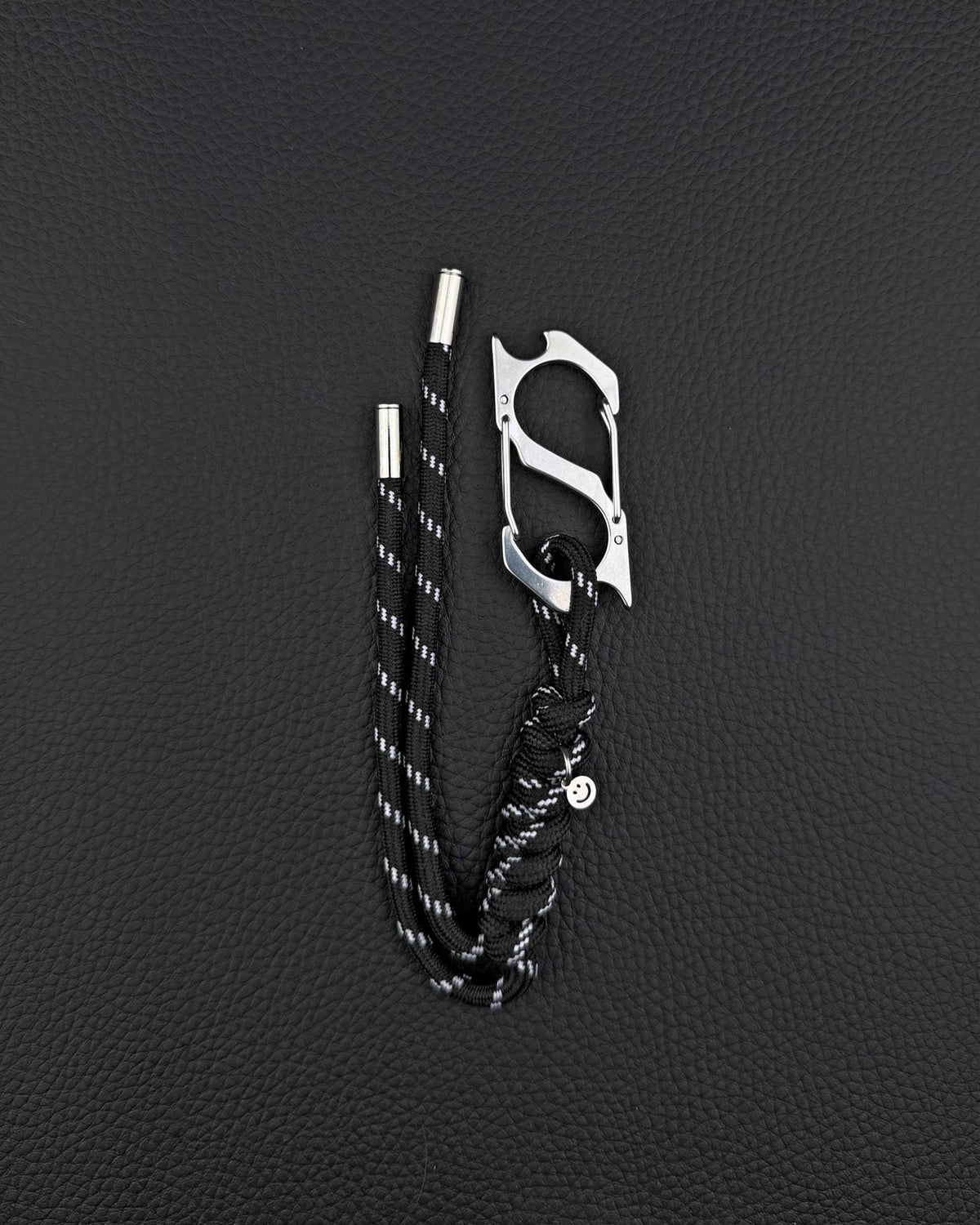 A black paracord keyring featuring a silver-toned stainless steel metal carabiner with an integrated bottle opener. The cord is woven with reflective white threads and finished with two silver metal aglets at the ends and a small circular charm. cyber sigilism style