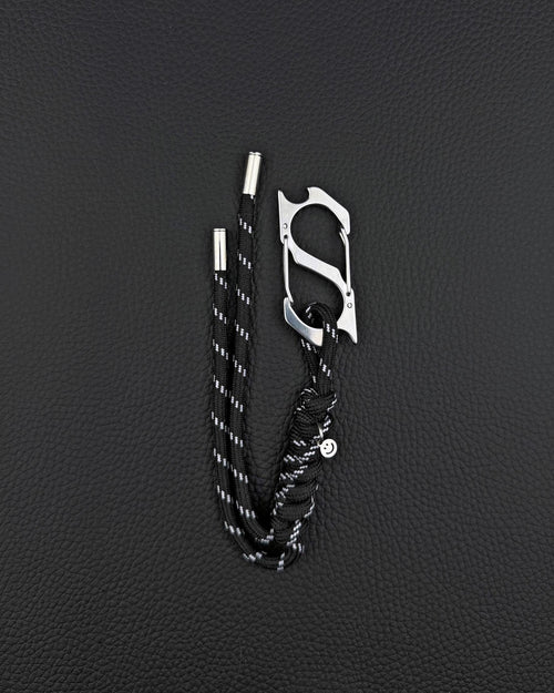 A black paracord keyring featuring a silver-toned stainless steel metal carabiner with an integrated bottle opener. The cord is woven with reflective white threads and finished with two silver metal aglets at the ends and a small circular charm. cyber sigilism style
