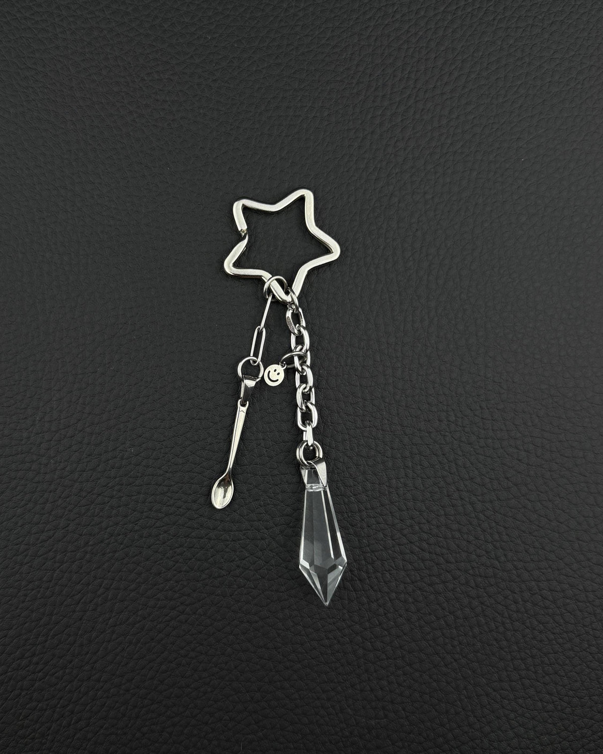 A silver stainless steel keyring featuring a star-shaped ring, a small stainless steel metal spoon charm, a tiny circular smiley face charm, and a clear, faceted, diamond-shaped glass crystal pendant hanging from a short metal chain link.