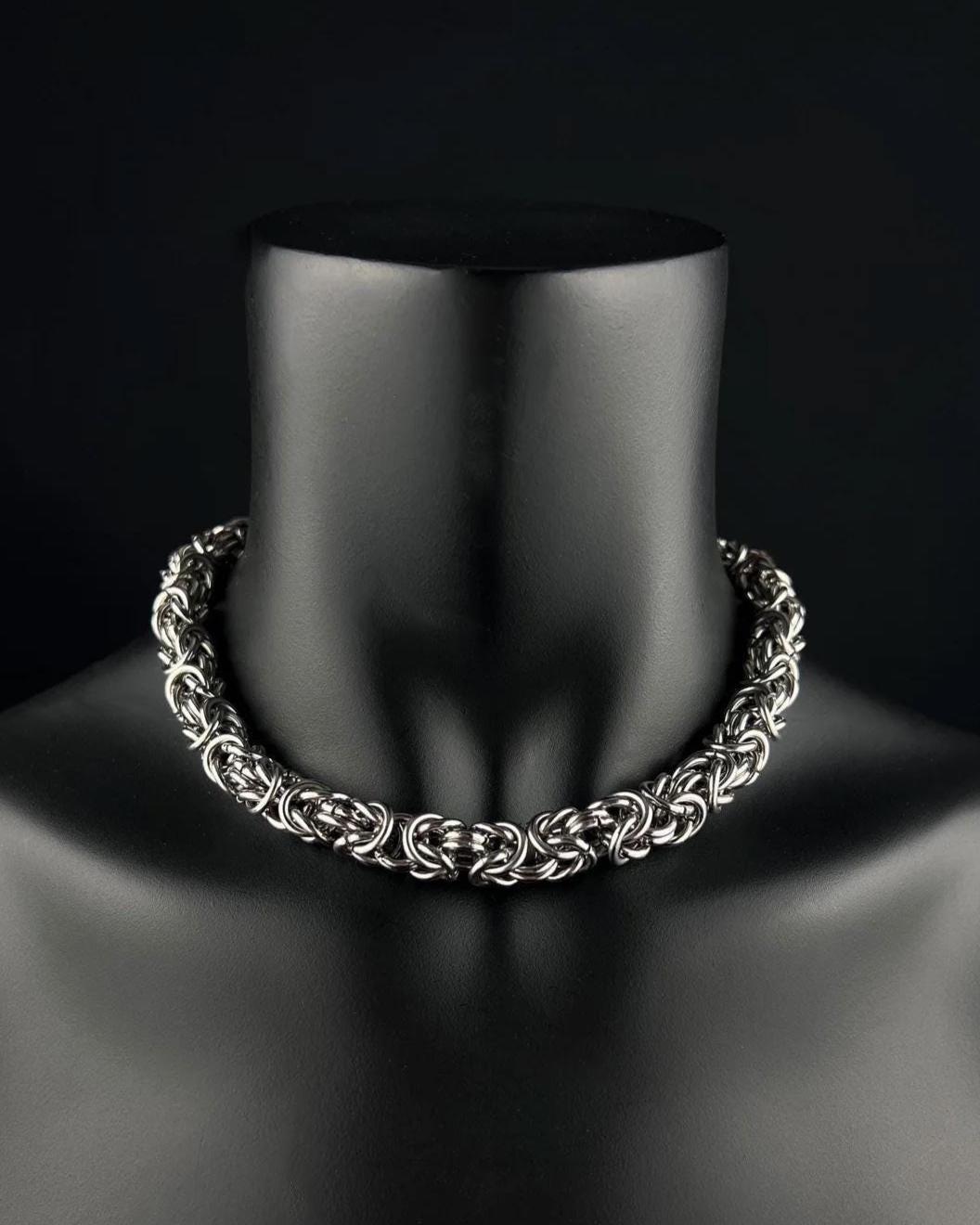 A stainless steel silver Byzantine chainmaille chunky chain necklace on a black mannequin. The necklace is made of interwoven metal rings, creating a thick, textured appearance. The background is a solid black.