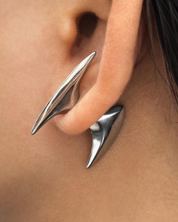 A silver stainless steel ear cuff featuring two distinct, smooth, pointed, and curved metal spikes. The larger, elongated spike rests along the upper outer edge of the ear, while the smaller, tapered spike sits below the earlobe, creating a sleek look.